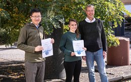 Two young researchers stand in front of trees holding their certificates, next to them stands Ludwig Stroink, who awarded the certificates.