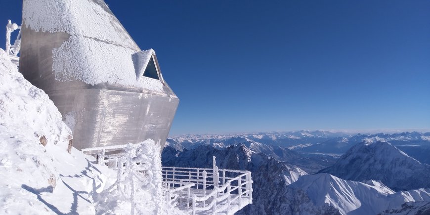 Ein futuristisch aussehender silberner Bau mit Balkon, über den beschneiten Gipfeln der Alpen.