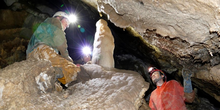 Zwei Menschen knien und stehen mit Helm und Stirnlampe in einer engen Tropfsteinhöhle