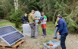 Fünf Menschen stehen um ein Bohrloch, das mit Beton abgeschlossen ist. Ein Solarmodul liefert Strom für Messgeräte. Im Hintergrund: Wald.