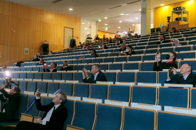 View up into the lecture hall: people sit and watch, some film the screen with their smartphones.