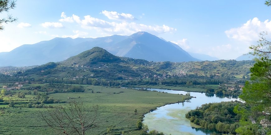 A green landscape. Middle and background: mountains. A river meanders in the foreground.