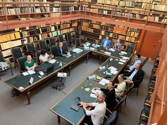 People sit at a U-shaped table in the historic library with papers in front of them and look into the camera.