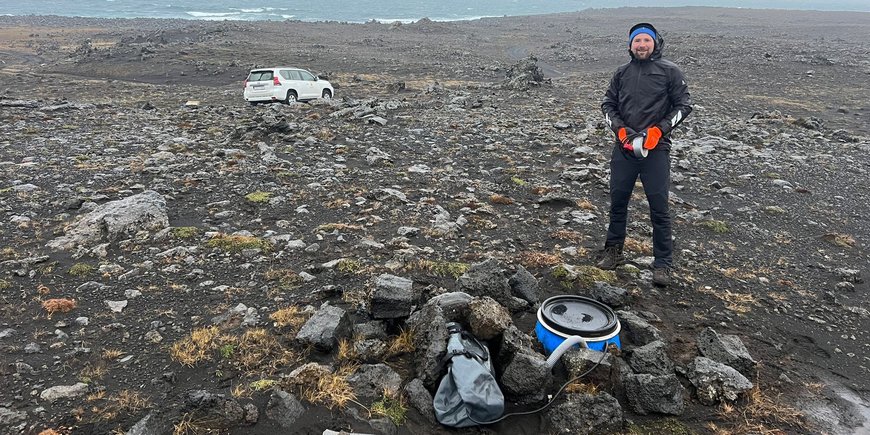 In the foreground Marius Isken and the seismometer, you don't recognise much of it, just a blue barrel embedded in the ground and several cables and hoses, in the background an off-road vehicle and the sea