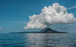 Insel-Vulkan mit Aschewolke, im Vordergrund ist ein ruhiges Meer zu sehen