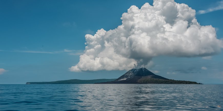 [Translate to English:] Insel-Vulkan mit Aschewolke, im Vordergrund ist ein ruhiges Meer zu sehen
