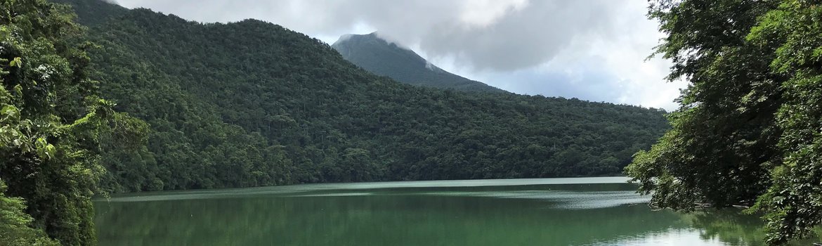 A green lake surrounded by green forested mountains, a volcano in the background.