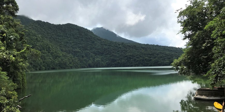 A green lake surrounded by green forested mountains, a volcano in the background.