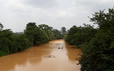 Brownish discoloured river meanders through green landscape with trees. Some people are standing in the river.