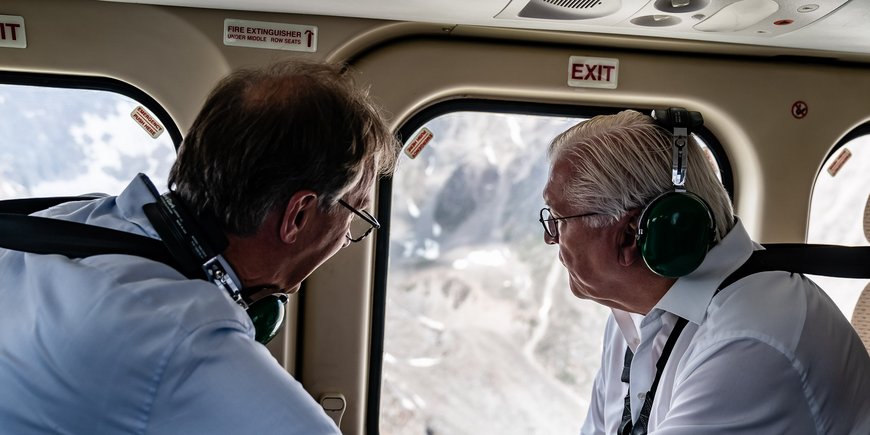 Two men sit opposite each other in an aeroplane and look out of the window at a mountain landscape.