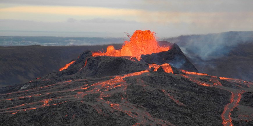 Dark grey, flat volcanic landscape, in the middle red-hot lava gushes out of a crater and runs down the slopes.