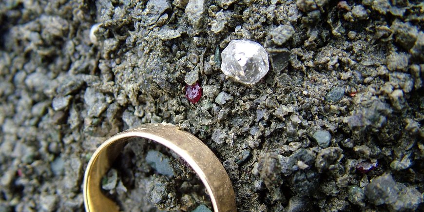 Ein Diamant, etwa so groß wie ein kleiner Fingernagel, in zerklüftetem Gestein. Zum Vergleich darunter: ein goldener Ehering.