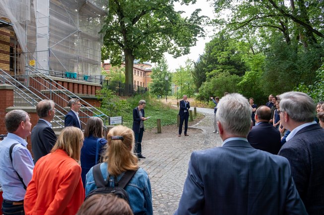 In front of a building, a group of people stand in a semicircle and listen to the speech of a person in the middle.