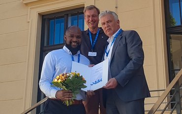 Three people are standing on a staircase in front of a yellow house: on the right, the award winner with a bouquet of flowers in his hand. He is being presented with the certificate.