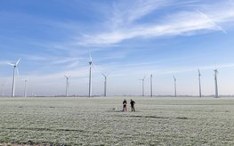 A few wind turbines on flat land. Two people are standing in front of them on the frosted field. Blue sky with hazy clouds.