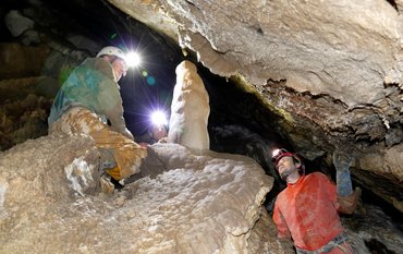 Two people kneel and stand with helmet and headlamp in a narrow stalactite cave