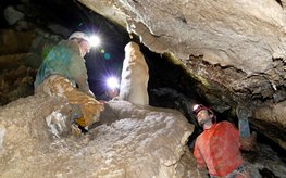 Two people kneel and stand with helmet and headlamp in a narrow stalactite cave