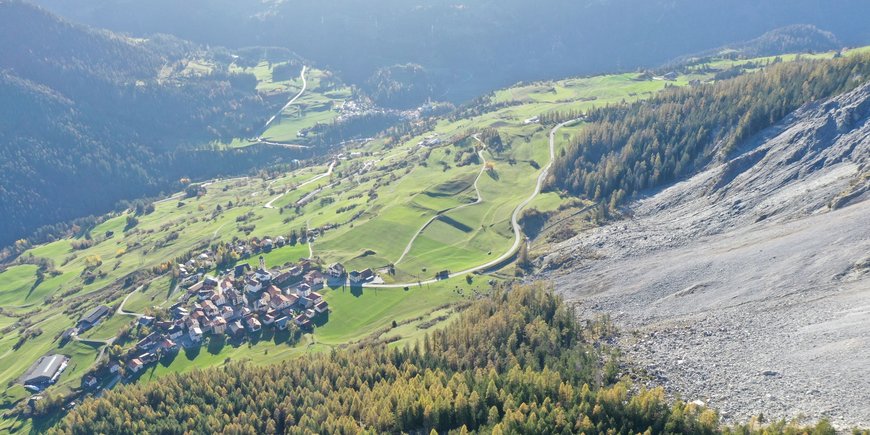 Blick am Geröllfeld entlang hinunter ins grüne Tal auf das bedrohte Dorf