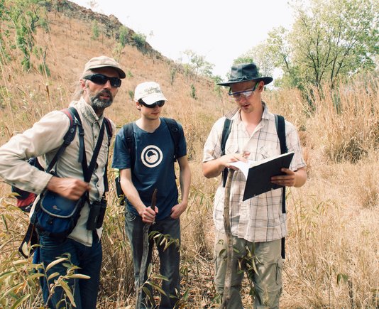 Close-up of three men walking through a mountainous, dry grassy landscape with hats and backpacks.
