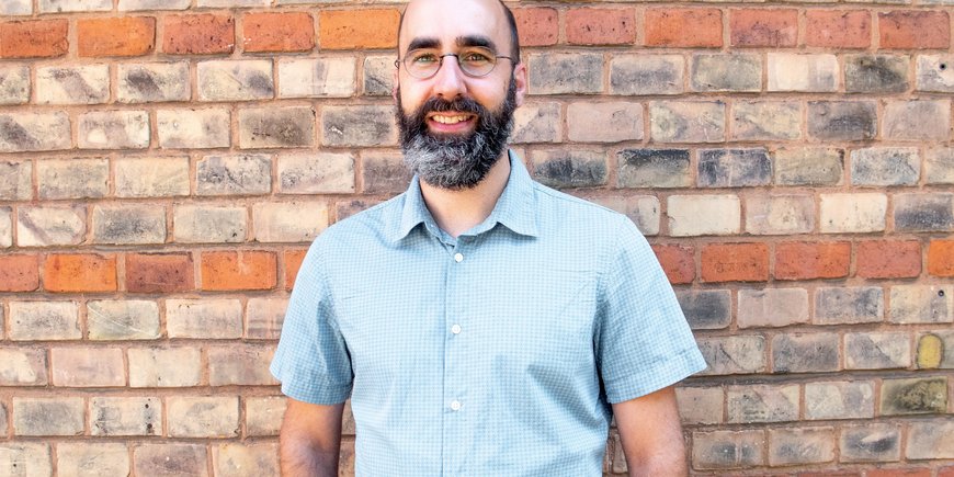 Christopher Kyba, bald man with glasses and beard, in short-sleeved blue shirt in front of brick wall