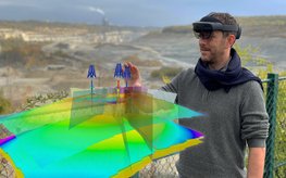 Eine Person steht mit aufgesetzter VR-Brille im Gelände, vor ihm ein digitales Geländemodell, mit dem er gerade interagiert.