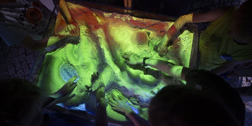 Several hands touch a modelled landscape with "mountains" and "valleys". Contour lines and different colours depending on the altitude are projected onto it.