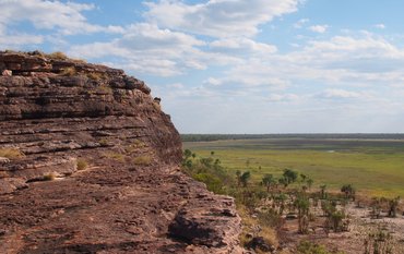 Landscape in northern Australia: Dark brown, layered rocks in the foreground on the left, view of a wide grassy landscape on the right.