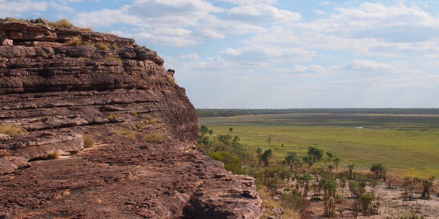 Landscape in northern Australia: Dark brown, layered rocks in the foreground on the left, view of a wide grassy landscape on the right.