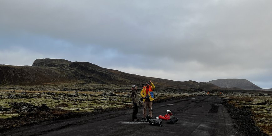 two people with luggage on a dirt track in Iceland, dark ground, mountains/rocks in the background