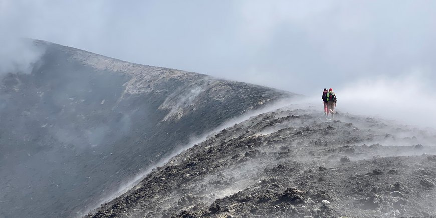 Zwei Personen/Forscher:innen die auf einem Kamm des Vulkan laufen, man sieht sie aus der Ferne und von hinten