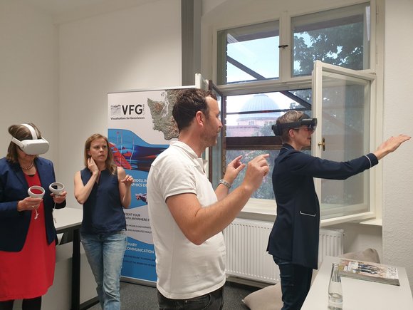 Two women wear VR/AR glasses and stretch out their arms. Two people stand in the middle and explain the technology to them.