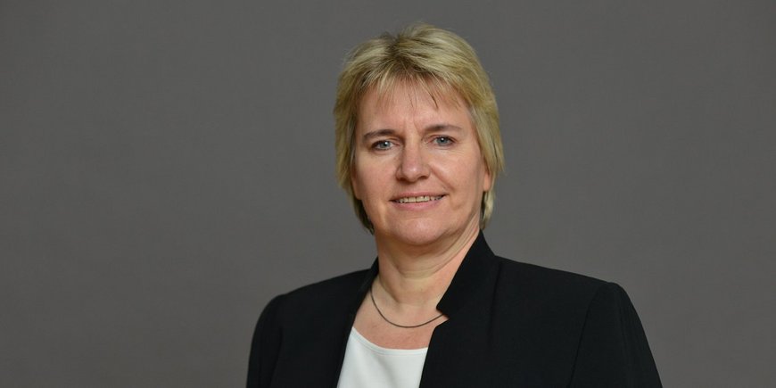 Portrait of a woman with short blond hair, black jacket over a white shirt against a dark grey background.