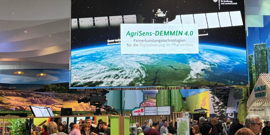 Großbildschirm mit Schriftzug Agrisens und einem Satelliten, aufgehangen in einer Halle auf der Landwirtschaftsmesse "Grüne Woche."