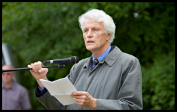 A man with grey hair and wearing a grey coat stands in front of green bushes and speaks into a microphone.