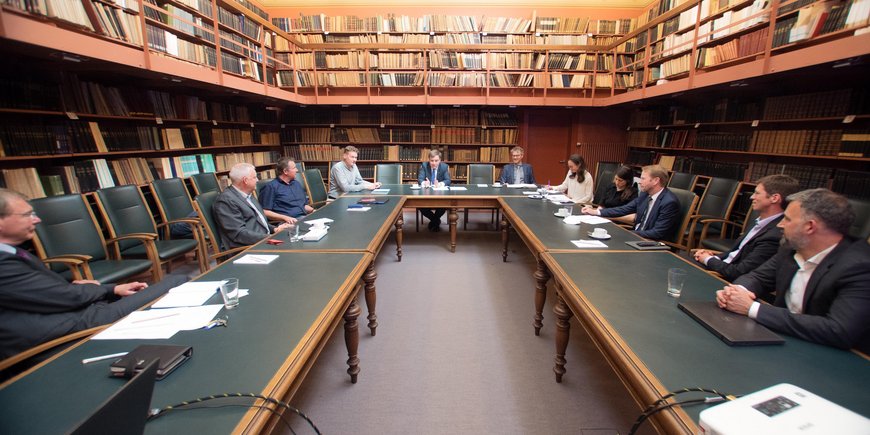 In the historic library, people sit around a u-shaped table and discuss, surrounded by books on the walls.