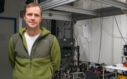 Sergey Lobanov steht mit grüner Vlies-Jacke in seinem Labor neben einem Tisch, auf dem eine Laser-Apparatur aufgebaut ist.