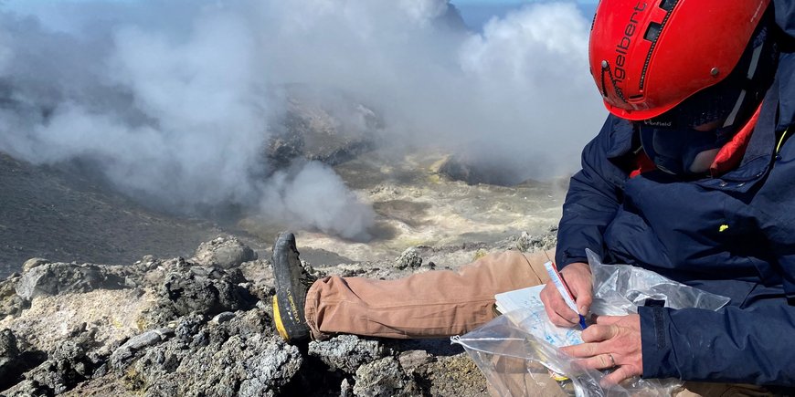 [Translate to English:] Forscher auf dem Vulkan Ätna mit Notizblock schreibend, im Hintergrund Aschewolken des Vulkans