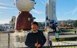 Ein junger Mann mit Brille steht lächelnd in einem Park vor einem Raumfahrzeug des Typs Sojus und einer Ariane-Weltraumrakete. Am blauen Himmel sind lange weiße Wolken und Kondensstreifen.