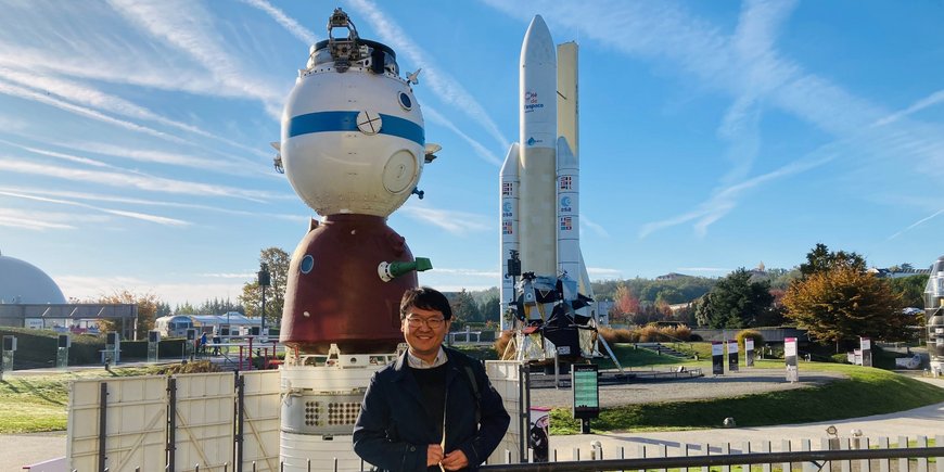 A young man with glasses stands smiling in a park in front of a Soyuz spacecraft and an Ariane space rocket. There are long white clouds and vapour trails in the blue sky.