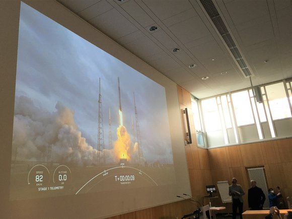 The screen in the lecture hall shows the rocket taking off.