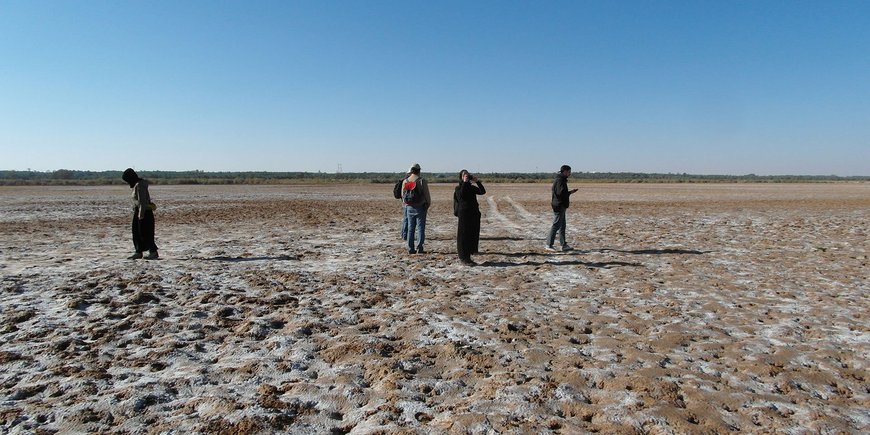 The barren, brown plain of the dried-up lake, on it a few people in long robes.
