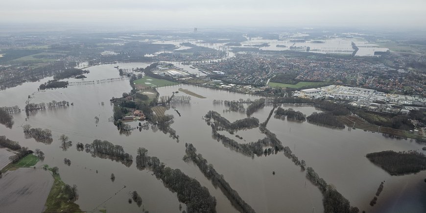 Bild aus dem Flugzeug von einer platten Landschaft, die großflächig überflutet ist: Felder, Straßen und Teile von Ortschaften stehen unter Wasser.