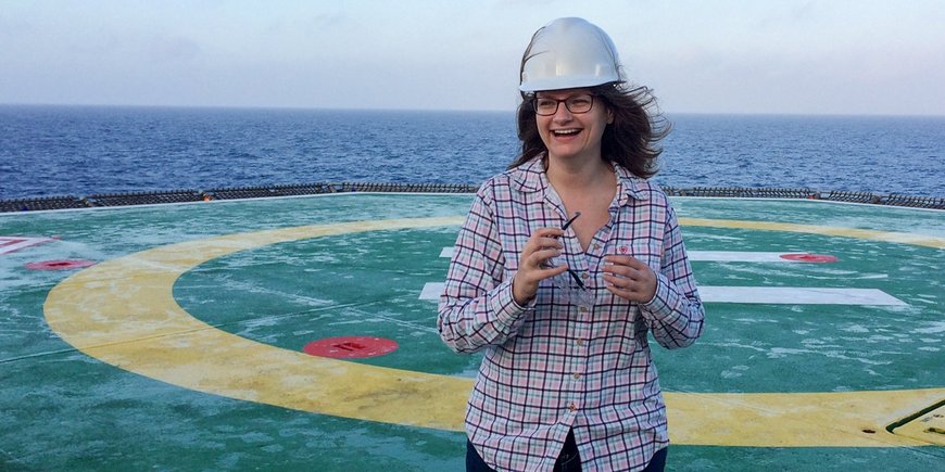 Eine junge Frau steht mit Sicherheitshelm auf einem Forschungsschiff.