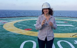 A young woman stands on a research vessel wearing a safety helmet.