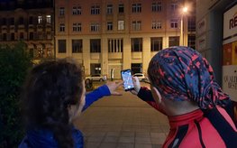 Zwei Menschen stehen nachts in einer Straße und halten ein Smartphone vor sich, um die Beleuchtung mit einer App zu registrieren.