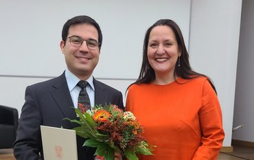 Portrait of Ugur Öztürk in suit and tie with certificate and bouquet of flowers. On the right side is Manja Schüle in a red dress.