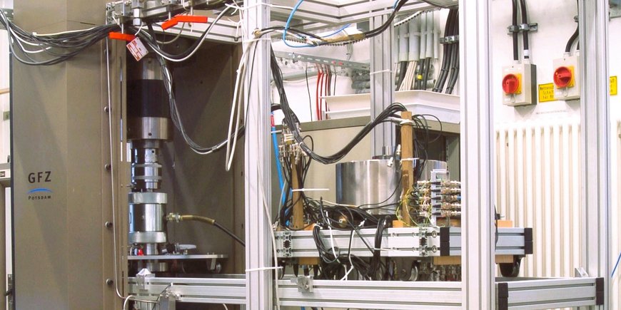Scene in the geomechanical high-pressure laboratory: metal frame with metal containers, cables, measuring instruments and a press.