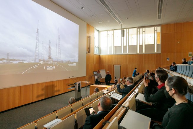 On the screen, the rocket shortly before launch. In the auditorium, two people comment.