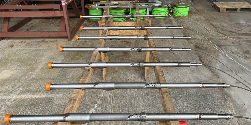 11 thin metal tubes, the borehole seismometers, lie lined up on a wooden rack above a concrete floor. Cables in the background.