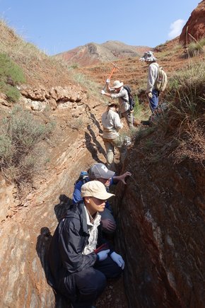 Several people stand in a trench in the dry mountainous area and take soil and rock samples.